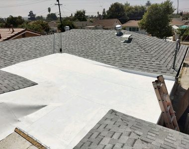 A flat white roof section surrounded by sloped gray shingles, with a ladder propped up against the edge and rooftops in the background.