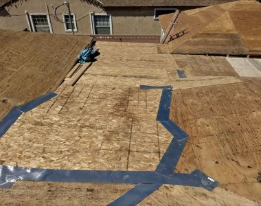 Roofing underlayment with plywood sheets, tools, and blue tape on a sloped roof, under bright sunlight.
