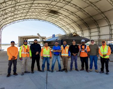 A group of men in orange vests stands in front of a fighter jet, showcasing teamwork and aviation expertise.