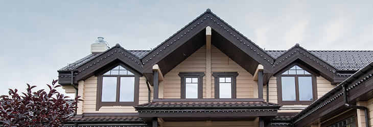 Elegant house exterior with a peaked roof, multiple windows, and decorative eaves against a cloudy sky. Bright foliage in the foreground.